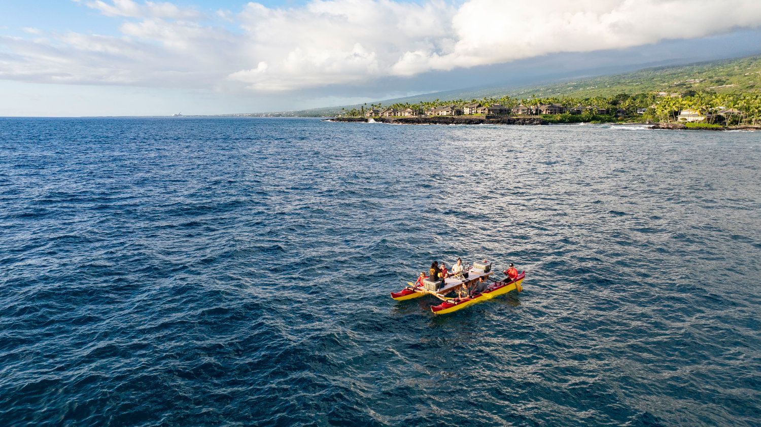 Ocean view with people in a yellow outrigger canoe. Lush green shoreline with clouds in the distance.