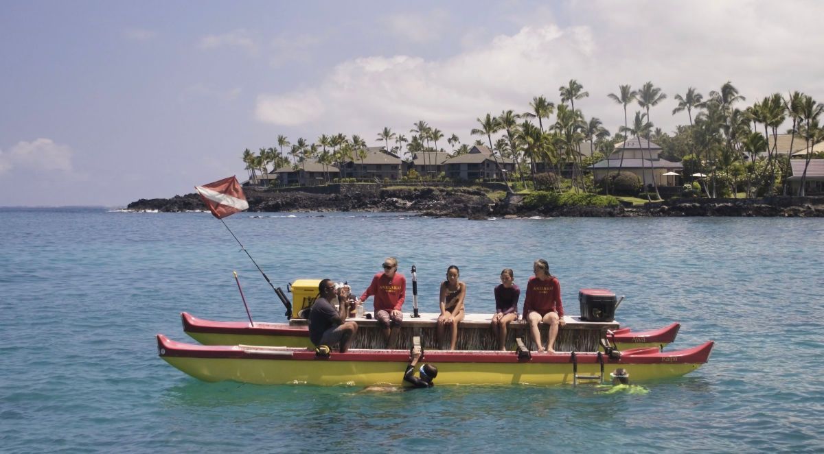 Yellow outrigger boat with people, diver down flag, in ocean near shore with buildings and palm trees.