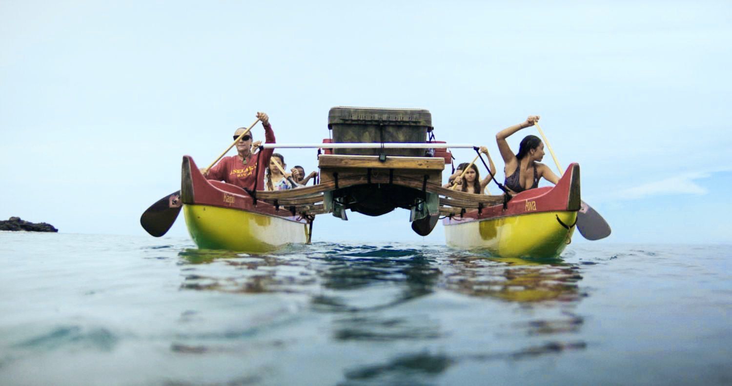 People paddling a catamaran with a cargo box on the ocean.