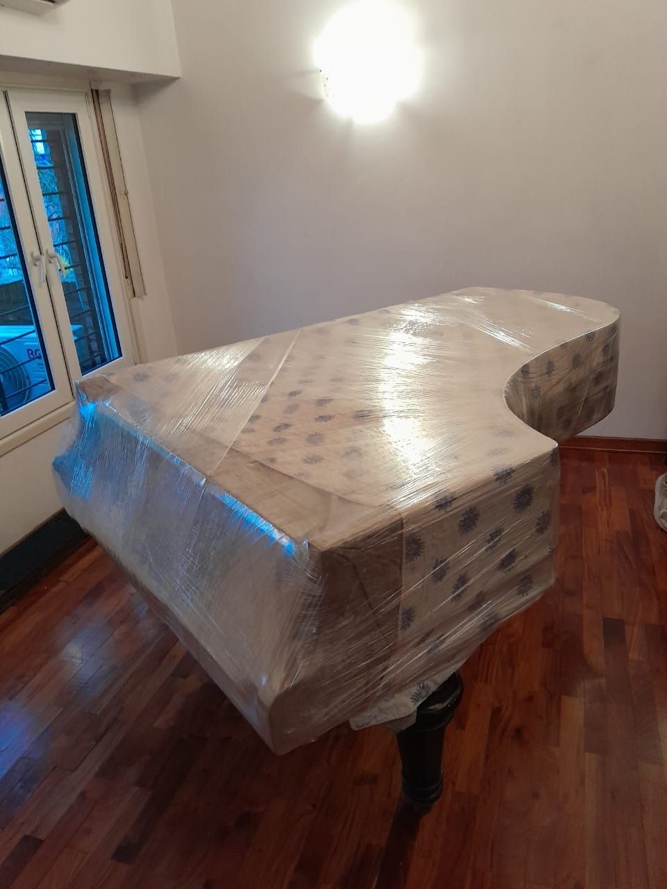 Piano envuelto en film plástico, listo para transportar, en una habitación con suelo de madera.