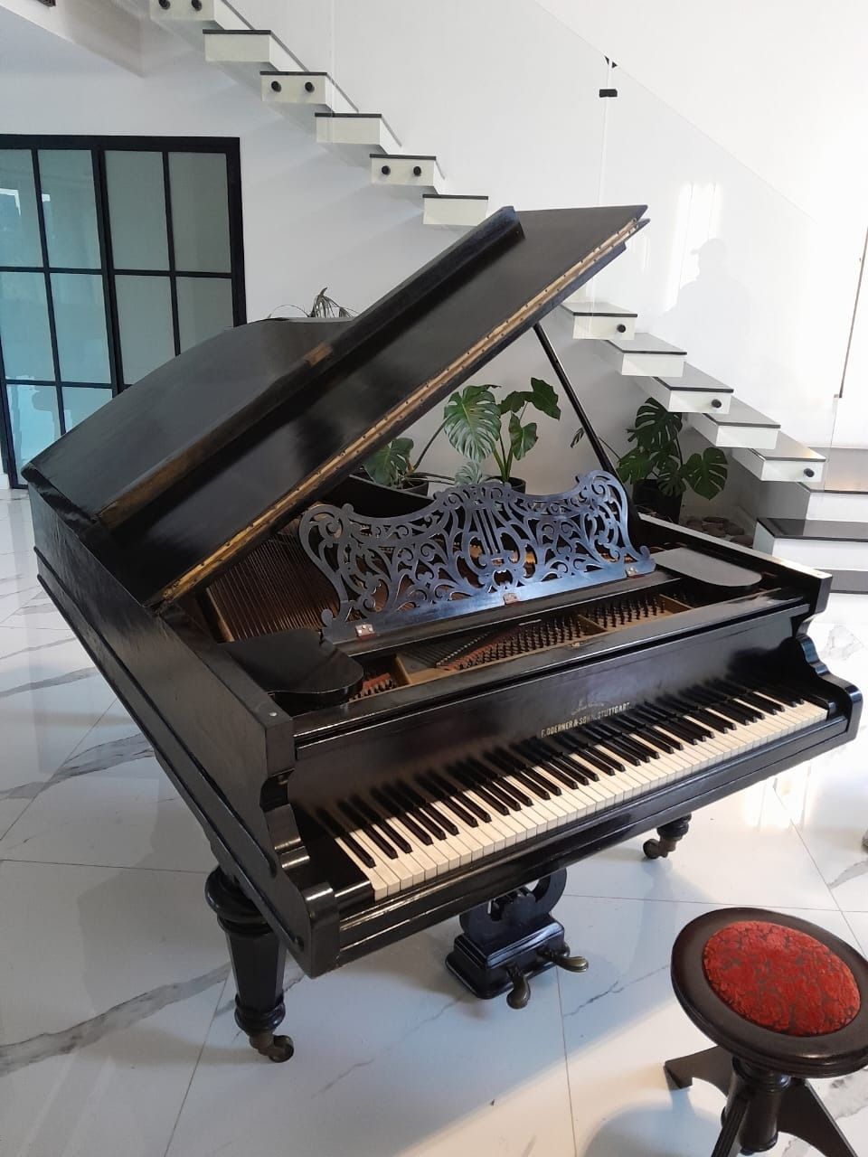 Piano de cola negro en una habitación luminosa con escalera y taburete rojo.