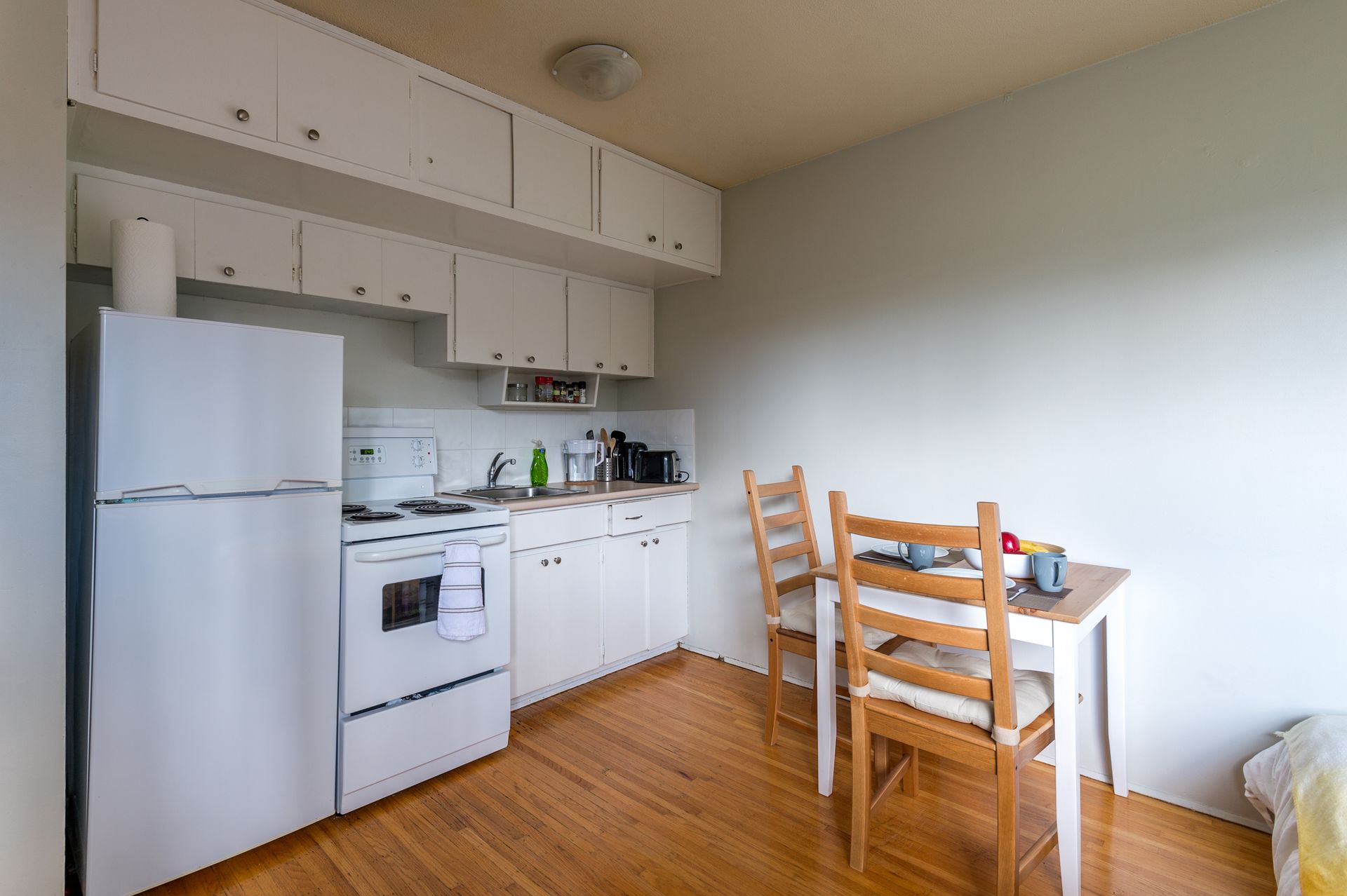 Small kitchen with white appliances, cabinets, and a wooden table with chairs by a bright wall.