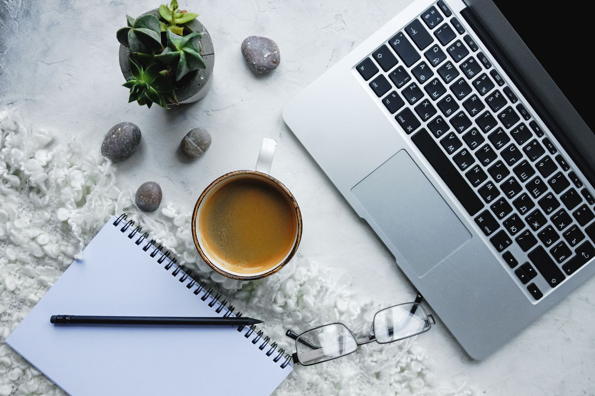 Desk with laptop, coffee, notebook, pen, glasses, and a small plant on a gray marble surface.