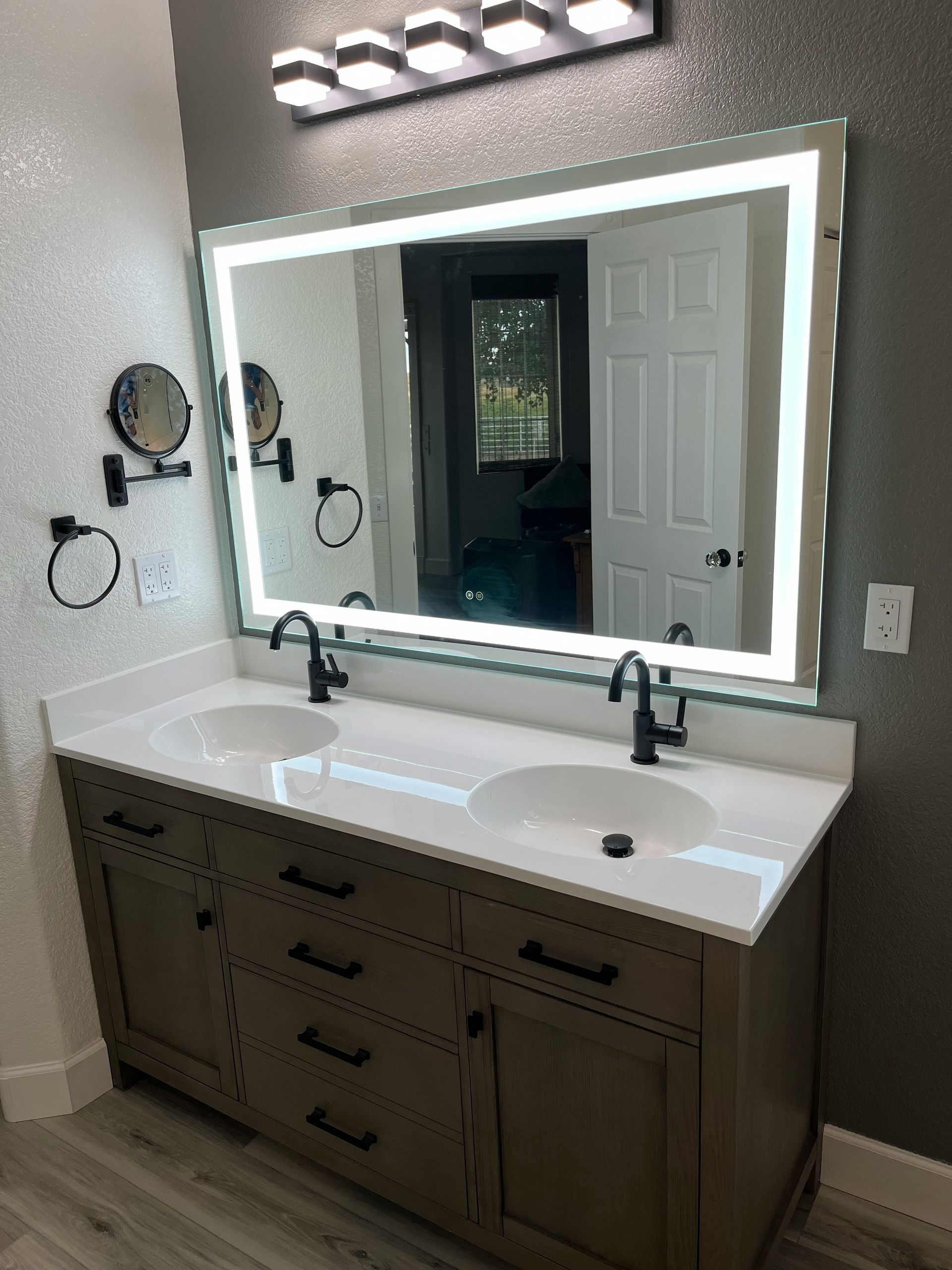 Double vanity bathroom remodel with gray shaker cabinets, white quartz countertop, dual undermount sinks, matte black faucets, LED backlit mirror, and wood-look LVP flooring — Handyman On Demand, Maryland