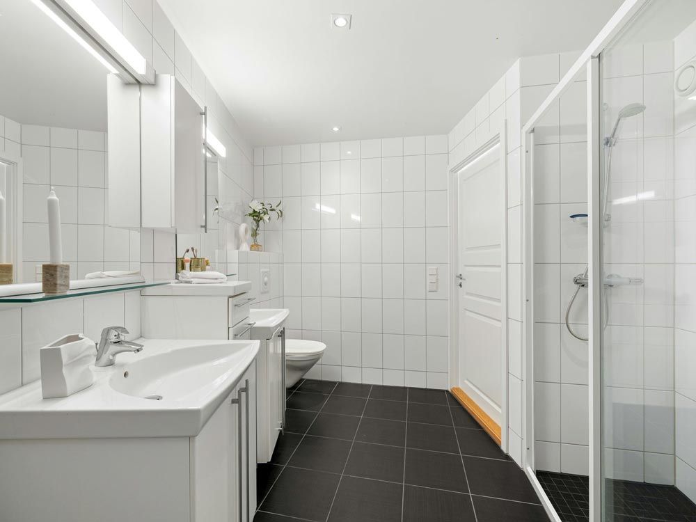 White-tiled bathroom with black floor, featuring a shower, two sinks, and a toilet