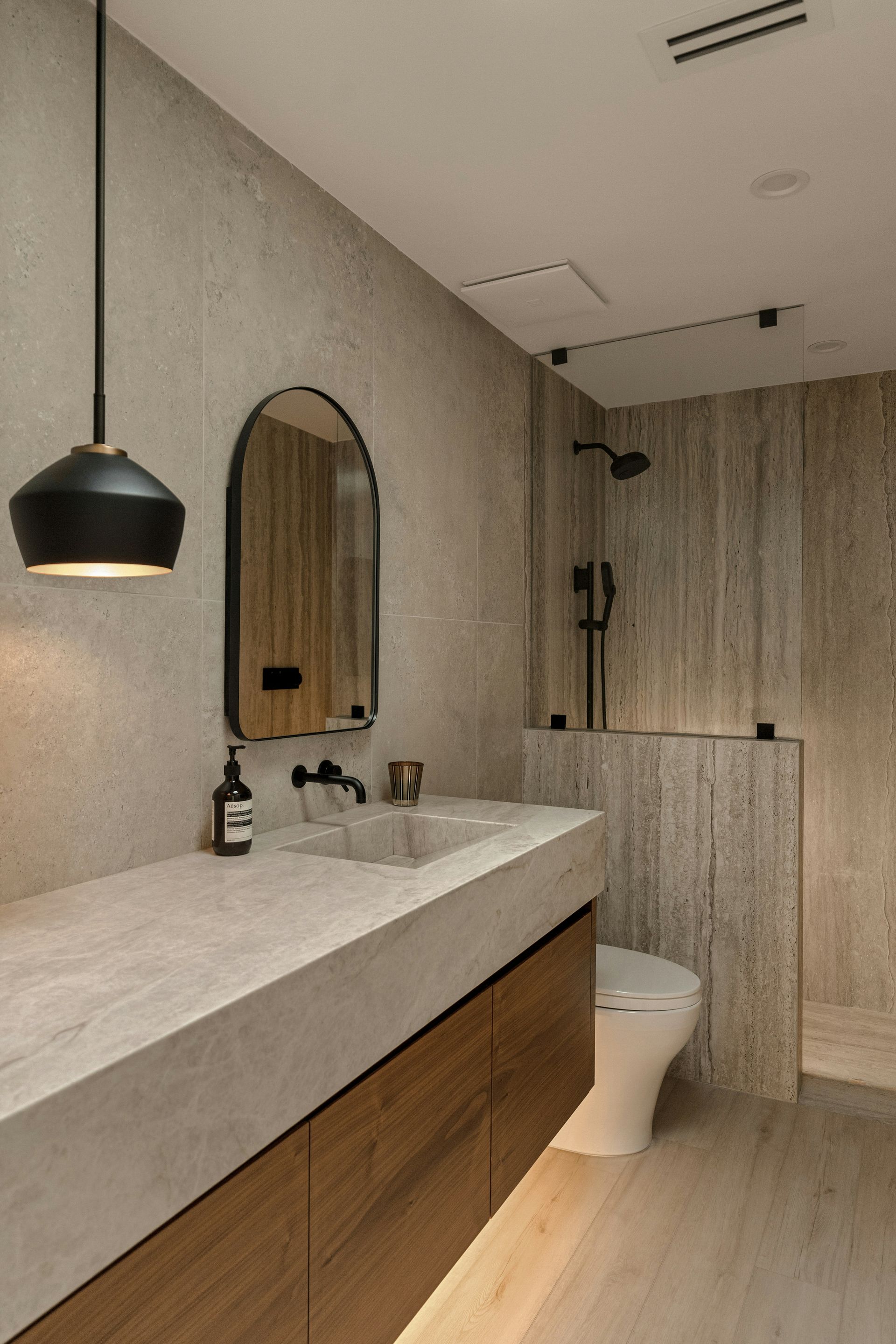 Modern luxury bathroom remodel with floating wood vanity, integrated marble countertop and sink, large format stone tile walls, open walk-in shower, arched black mirror, black pendant light, and matte black fixtures — Handyman On Demand, Maryland
