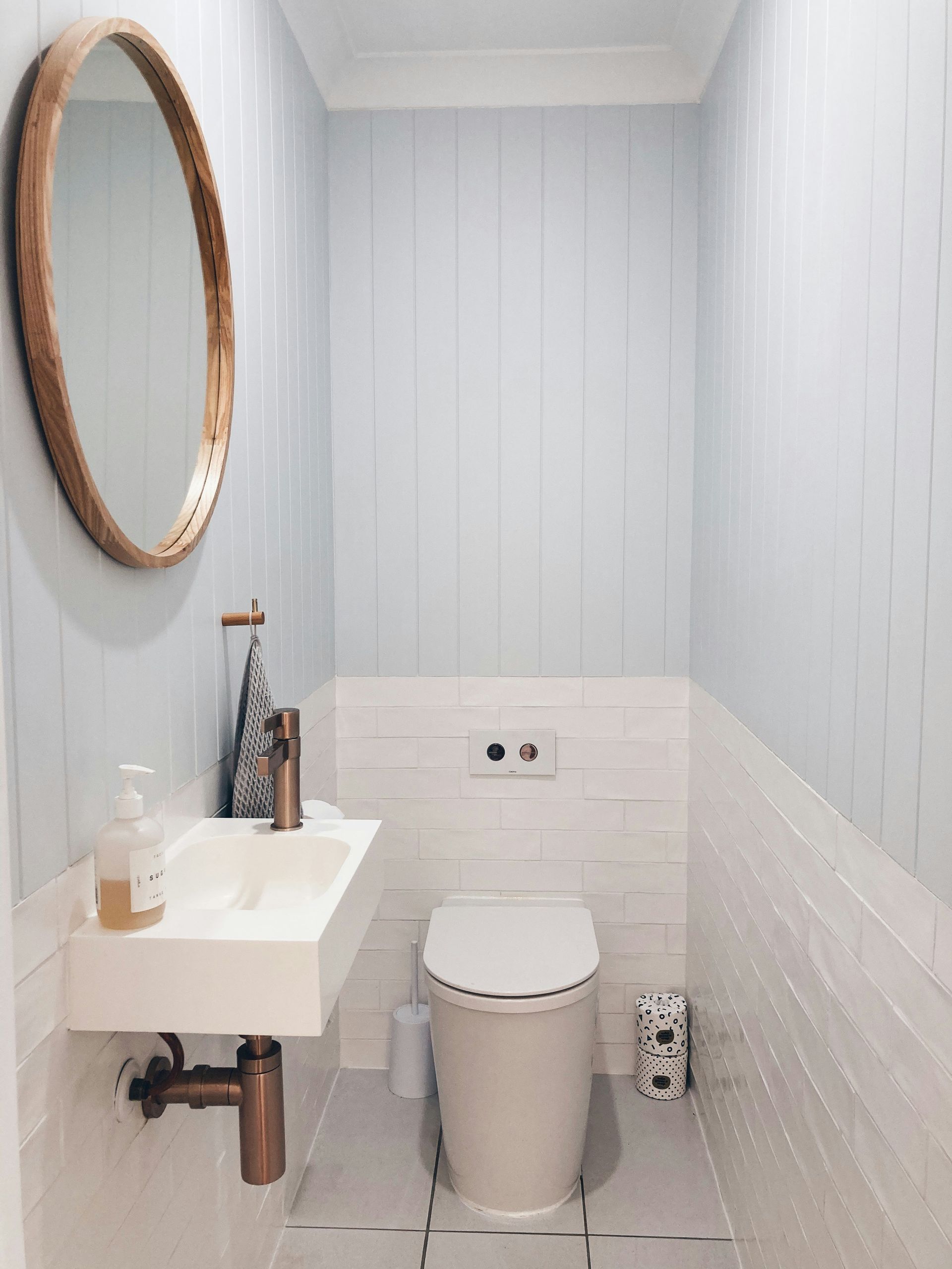 Powder room remodel with painted vertical shiplap walls, white subway tile wainscoting, wall-mounted rectangular sink, exposed bronze faucet and pipe, round wood-framed mirror, and wall-mounted toilet — Handyman On Demand, Maryland