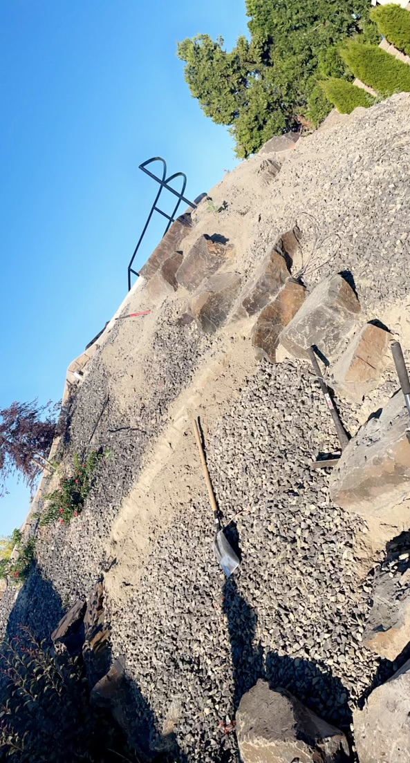 A steep, rocky incline features a stone staircase leading up to a simple black metal handrail against a blue sky.