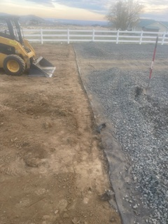 A yellow skid steer sits on a dirt lot next to a section of ground covered in gray gravel and landscape fabric.