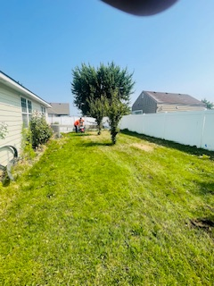 A person in an orange shirt mows the grass in a sunny backyard between a house and a white vinyl fence.