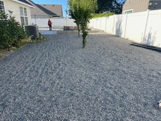 A gravel yard in a residential backyard, bordered by a white fence and the side of a house with a person standing nearby.