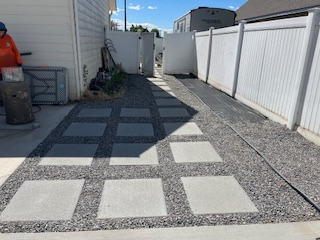 A backyard path featuring square concrete pavers set in dark gravel, leading toward a white fence and open gate.
