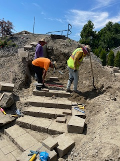 Three workers in high-visibility clothing install stone stairs on an outdoor hillside construction site.