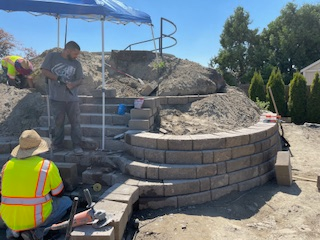 Workers build a curved stone retaining wall and stairs outdoors under a blue canopy on a sunny day.