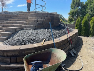 Construction of a tiered stone retaining wall and staircase with a wheelbarrow and shovels in the foreground.