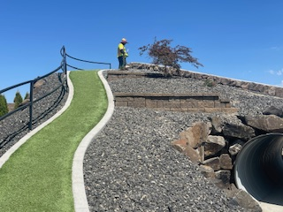 A worker stands on a rocky hillside next to a curved, green walking path and a large tunnel opening against a blue sky.