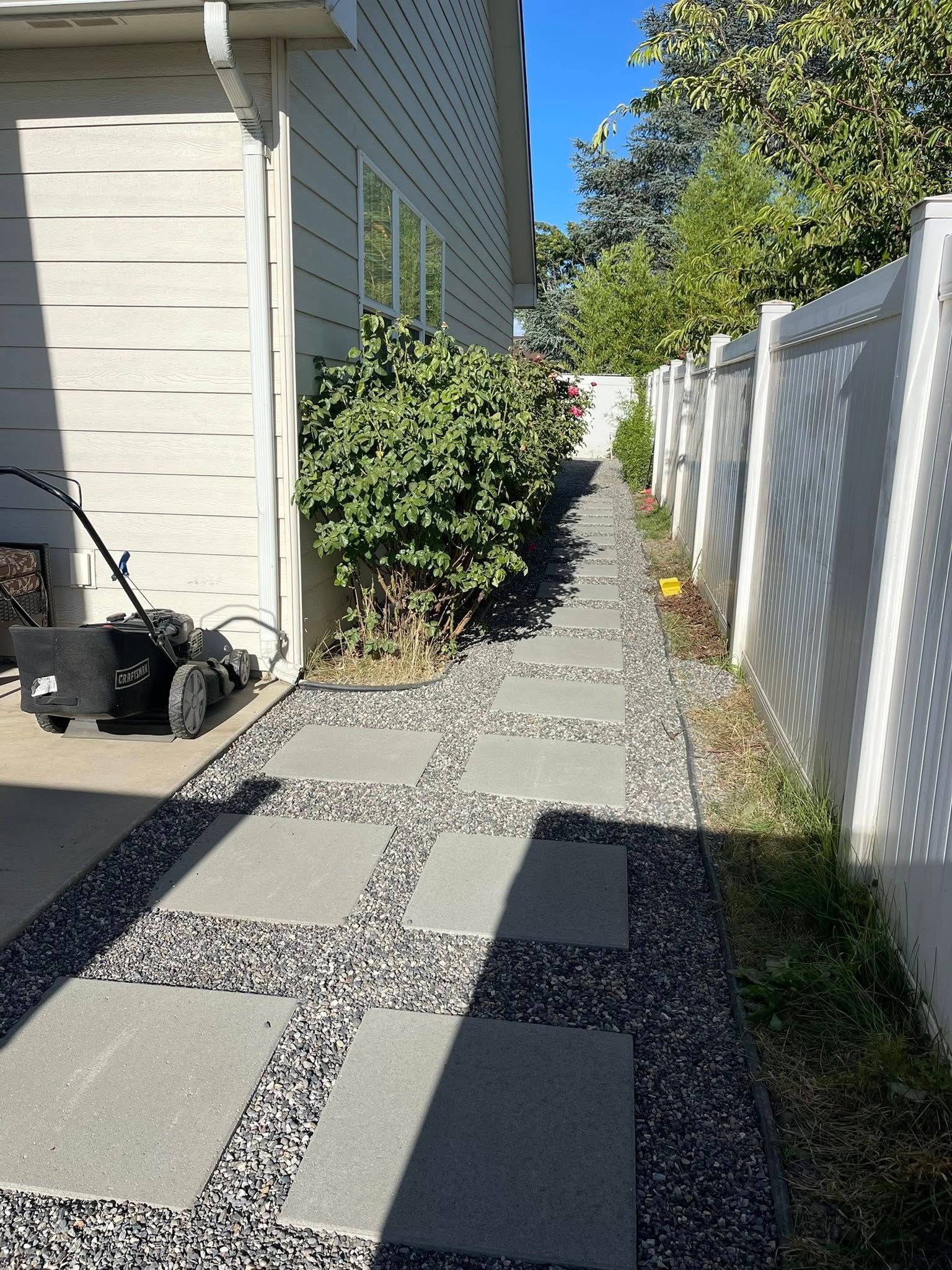 A path made of square concrete pavers set in dark gravel alongside a house wall and a white privacy fence.
