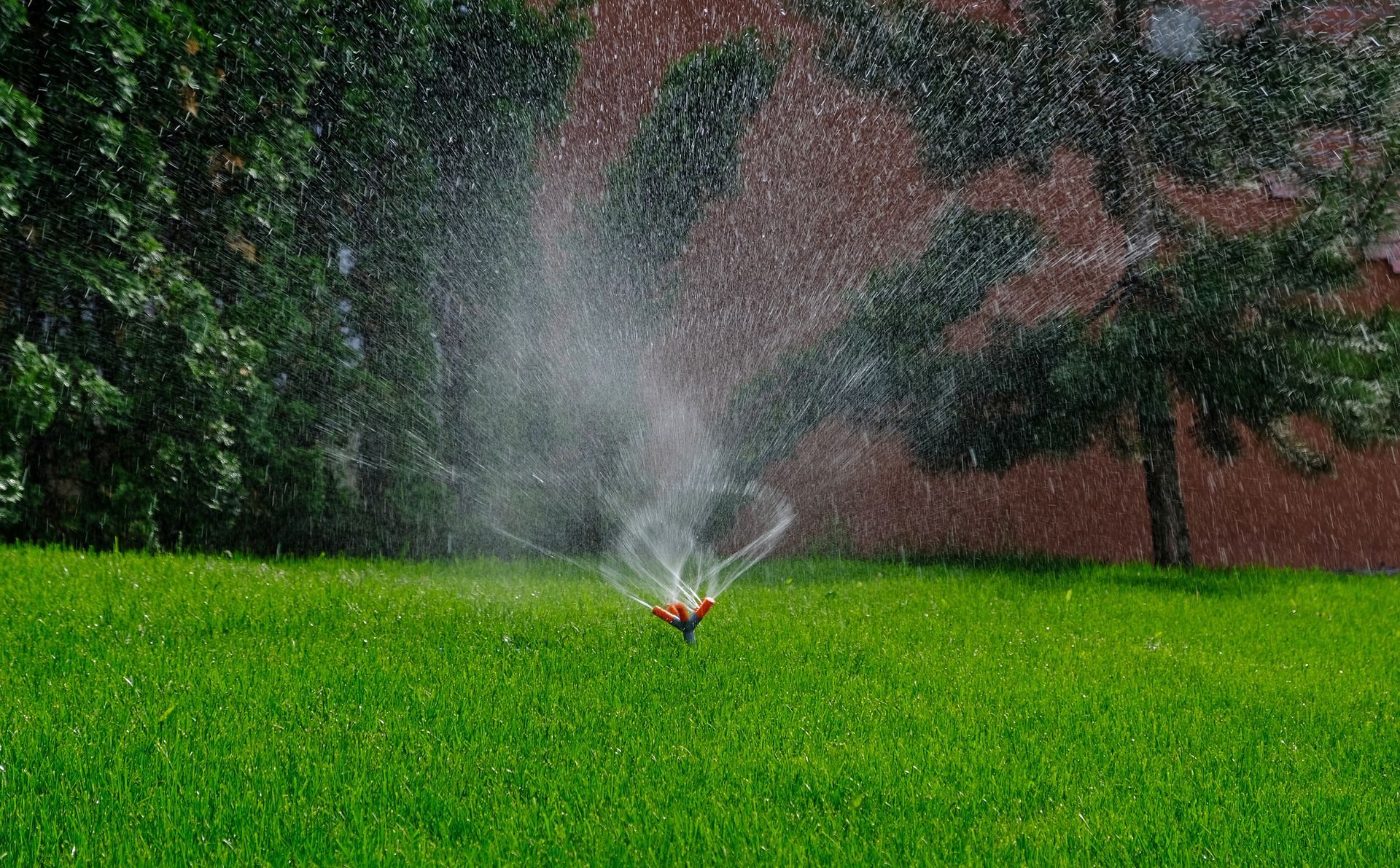 A sprinkler sprays water across a vibrant green lawn in front of a red brick building and trees.