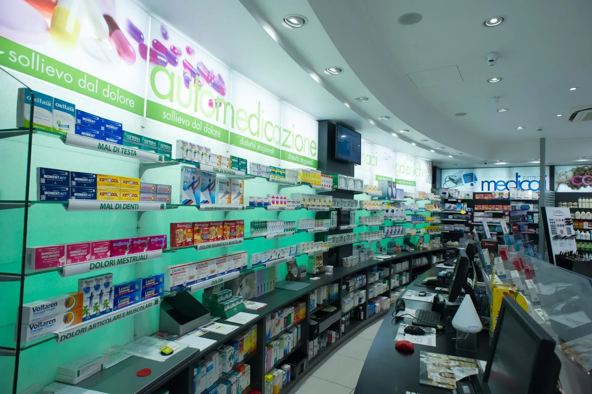 Interno di una farmacia con scaffali pieni di medicinali e bancone curvo. Design verde e bianco.