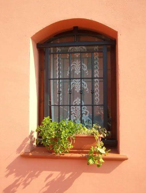 Finestra ad arco con inferriate nere, tenda di pizzo e una pianta verde in vaso su una parete intonacata color pesca.