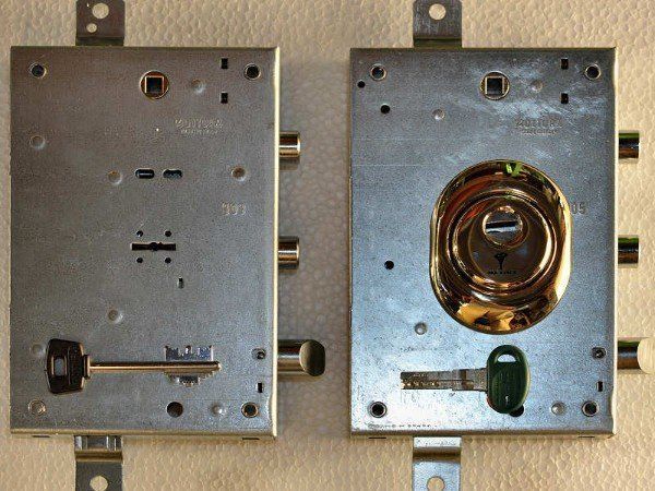 Due meccanismi di chiusura per porte in metallo affiancati, uno visto frontalmente e l'altro che mostra il chiavistello e il cilindro.