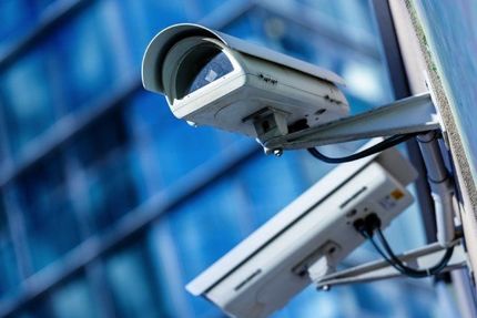 Due telecamere di sicurezza bianche montate su un edificio, inclinate verso l'esterno contro uno sfondo di vetro blu.