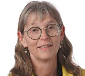 Kvinde med briller smiler til kameraet, iført en gul blazer og øreringe.