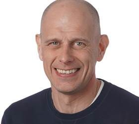 Skaldet mand smilende, iført en mørkeblå skjorte med rund hals, indendørs.