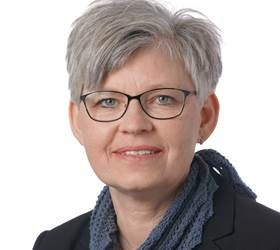 Kvinde med kort gråt hår og briller, iført en mørk blazer og blåt tørklæde, smilende.