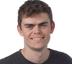 Mand med krøllet brunt hår smiler, iført en mørkegrå skjorte.