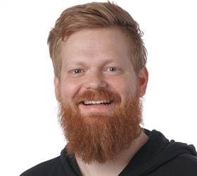 Mand med rødt hår og skæg smilende, iført en sort top mod en hvid baggrund.