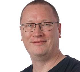 Mand med briller smilende, iført en mørk skjorte, mod en hvid baggrund.