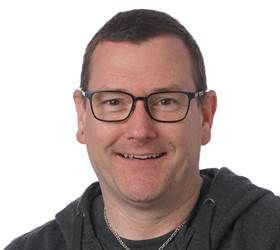 Mand med briller smilende, iført en mørk hættetrøje.