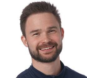 Smilende mand med kort brunt hår og skæg, iført en blå skjorte.