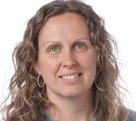 Kvinde med krøllet hår og blå øjne smiler til kameraet, iført hoop-øreringe.