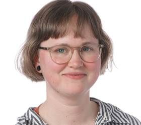 Kvinde med briller smiler; kort brunt hår, iført en stribet skjorte mod en hvid baggrund.