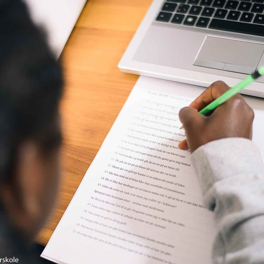 Person, der skriver med en grøn pen på et papir med tekst, bærbar computer til højre.