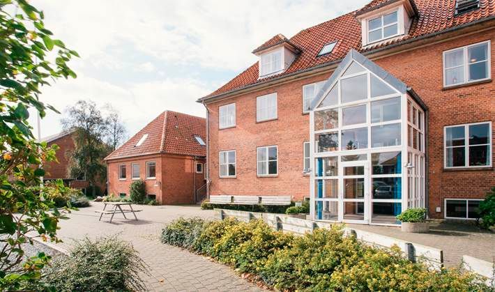 Rød murstensbygning med glasindgang, rødt tegltag og et brolagt udendørs område.