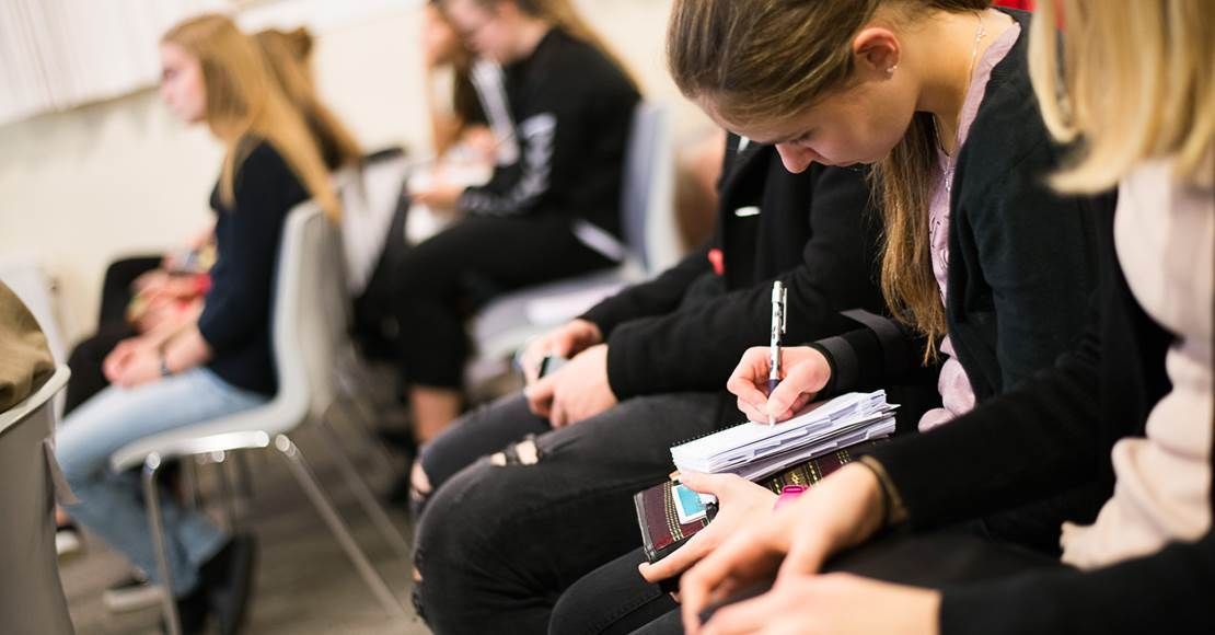 Folk sidder og tager noter i et klasseværelse. En person skriver med en pen i en lille notesbog.