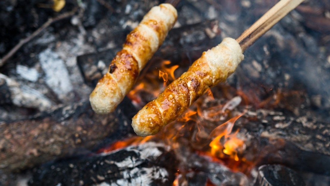 To pølser, der steges over et bål, spydt på træpinde.