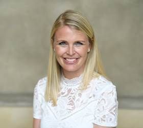 Blond kvinde smiler, iført en hvid blondetop. Lys baggrund.