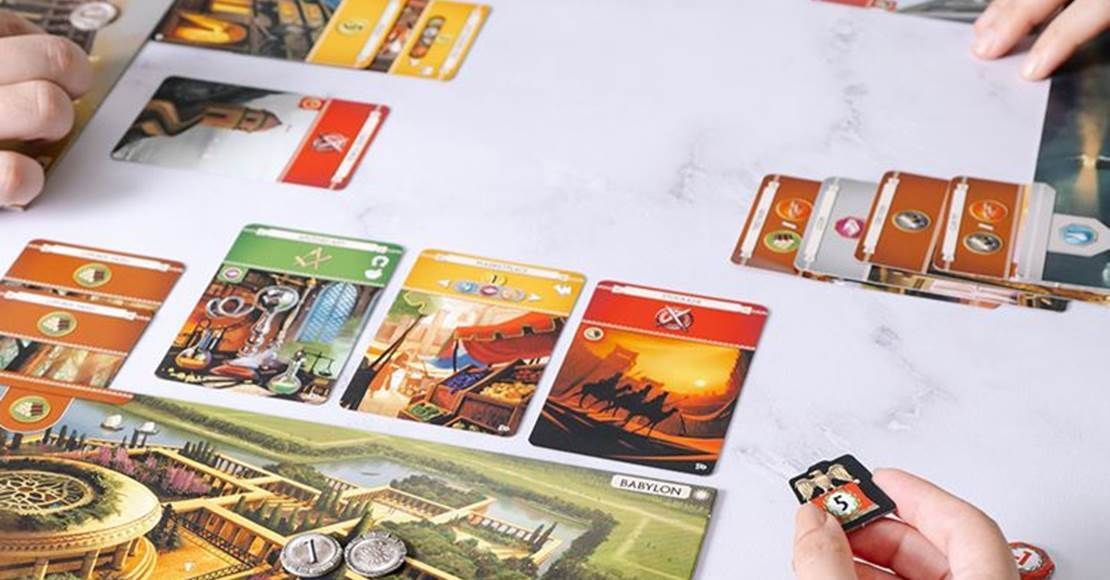 Brætspil i gang med kort, poletter og hænder på en hvid overflade.