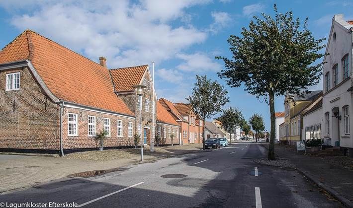 Gade i Løgumkloster by med murstens- og hvide bygninger, røde tegltage og træer. Solskinsdag.