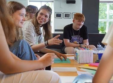 Gruppe af mennesker sidder omkring et bord med tuscher og papir, nogle smiler og taler. Indendørs.