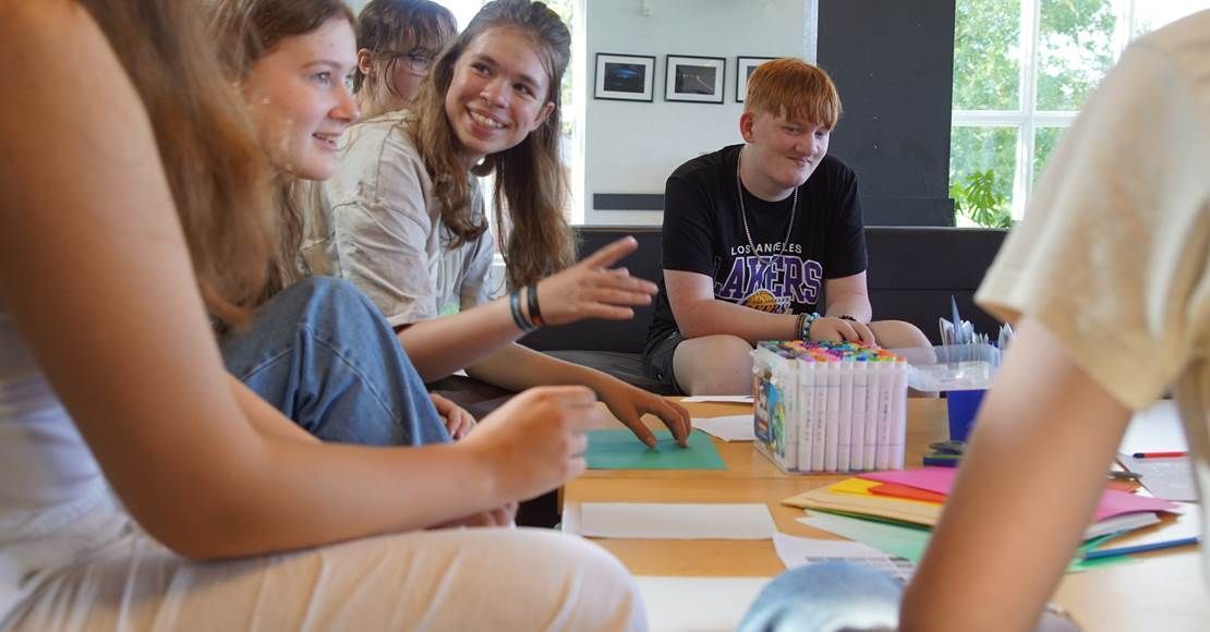 Gruppe af mennesker sidder omkring et bord, griner og taler i et godt oplyst rum. Farverigt papir og tuscher på bordet.