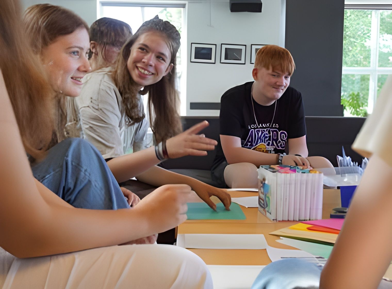Gruppe af mennesker sidder omkring et bord og tegner. Smiler, diskuterer, tuscher og farverigt papir synlige. Indendørs.
