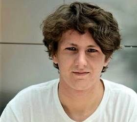 Person med krøllet brunt hår, iført en hvid t-shirt, smilende.