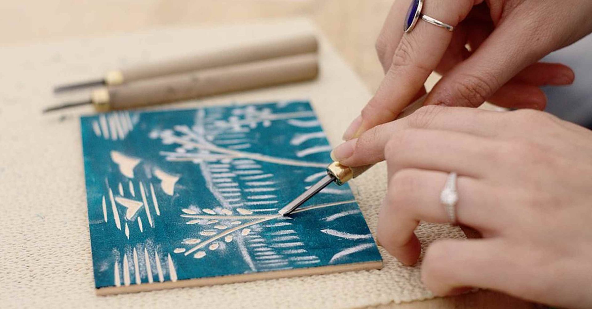 Hænder udskærer en blågrøn linoleumssnitblok med et lille værktøj, fugle og planter synlige.