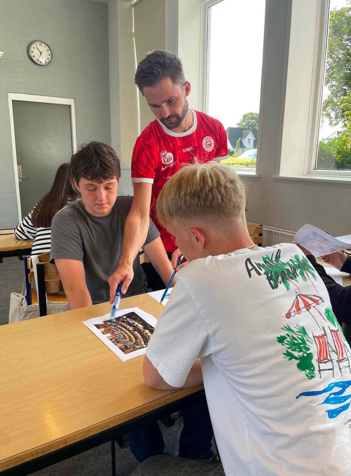 Tre personer undersøger et foto ved et bord. En mand i rød skjorte peger, mens to andre ser til. Klasseværelsesmiljø.