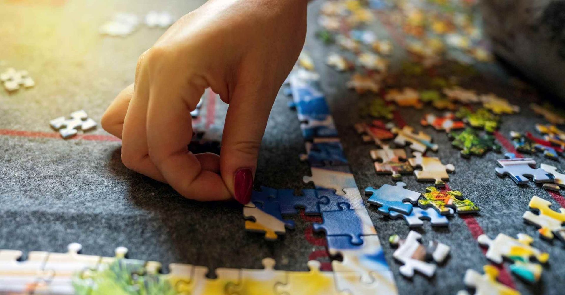 Hånd placerer en puslespilsbrik i et delvist færdiggjort puslespil; farverige brikker på en grå overflade.